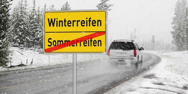 Schild „Winterreifen“ mit durchgestrichenem „Sommerreifen“ an einer verschneiten Straße
